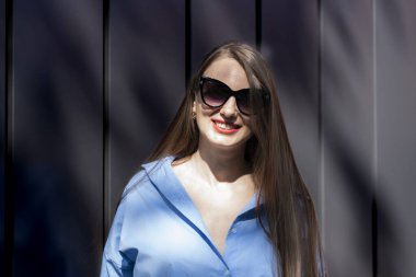 Güneş gözlüklü, uzun sarı saçlı bir kızın yaz portresi. Mavi gömlekli ve kot pantolonlu bir kız siyah bir duvarın karşısında duruyor. Yüksek kalite fotoğraf