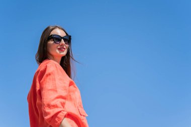 Uzun sarı saçlı bir kızın yaz portresi. Mavi gökyüzüne karşı turuncu elbiseli ve güneş gözlüklü kız. Yüksek kalite fotoğraf