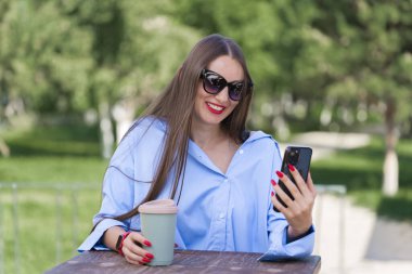 Uzun sarı saçlı bir kızın yaz portresi. Mavi tişörtlü ve güneş gözlüklü bir kız elinde bir akıllı telefon tutuyor ve video görüşmesiyle konuşuyor. Yüksek kalite fotoğraf