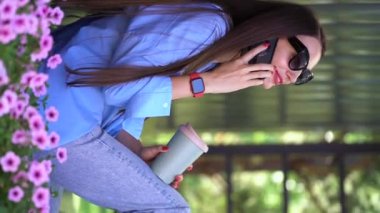 Uzun saçlı bir kız parkta bir bankta oturuyor, elinde termal bardağı tutuyor ve akıllı bir telefonla konuşuyor..