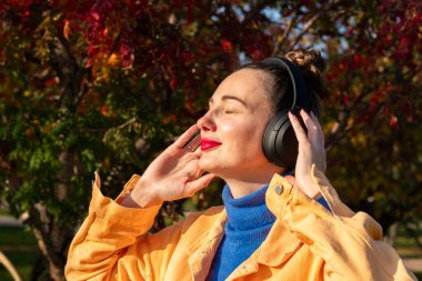 Kulaklıklı genç bir kadın müzik dinler ve sonbahar parkında yürür. Yüksek kalite fotoğraf