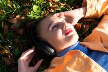 Genç bir kadın parktaki yeşil çimlere uzanıyor ve kulaklıklarıyla müzik dinlemekten hoşlanıyor. Yüksek kalite fotoğraf