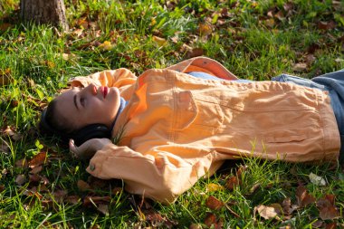 Genç bir kadın parktaki yeşil çimlere uzanıyor ve kulaklıklarıyla müzik dinlemekten hoşlanıyor. Yüksek kalite fotoğraf