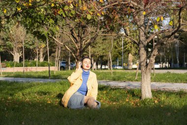 Genç bir kadın parktaki yeşil çimenlerde oturur ve kulaklıklarıyla müzik dinlemekten hoşlanır. Yüksek kalite fotoğraf