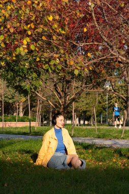 Genç bir kadın parktaki yeşil çimenlerde oturur ve kulaklıklarıyla müzik dinlemekten hoşlanır. Yüksek kalite fotoğraf