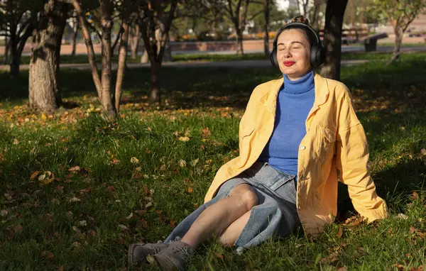 Genç bir kadın parktaki yeşil çimenlerde oturur ve kulaklıklarıyla müzik dinlemekten hoşlanır. Yüksek kalite fotoğraf