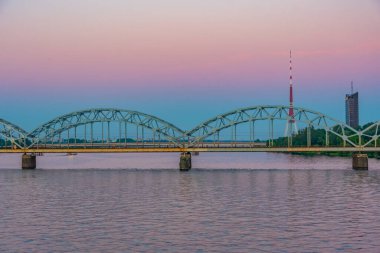 Riga 'daki radyo kulesinin günbatımı manzarası Letonya' daki çelik bir demiryolu köprüsünün arkasında..