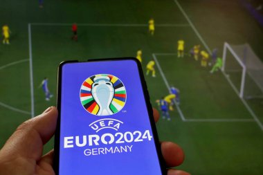 Bu resimde, UEFA Euro 2024 (2024 UEFA Avrupa Futbol Şampiyonası) logosu bir futbol maçının arka planında bir akıllı telefondan görülmektedir..