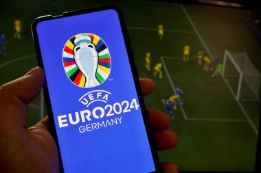 Bu resimde, UEFA Euro 2024 (2024 UEFA Avrupa Futbol Şampiyonası) logosu bir futbol maçının arka planında bir akıllı telefondan görülmektedir..