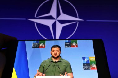 Viewing on a phone a live broadcast of Volodymyr Zelenskyy's speech against the background of the NATO flag during the 2023 NATO Summit in Vilnius, Lithuania, July 12, 202 clipart
