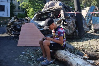 KYIV, UKRAINE - 20240709: Bir çocuk telefonda yıkılan bir apartmanın arka planına karşı oynuyor ve arabaları yakıyor.