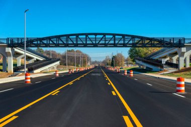 Newly-painted stripes and orange traffic barrels are seen on a freshly-blacktopped road under a pedestrian walkway bridge. clipart