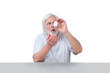 Horizontal shot of an elderly man sitting at a table examining his precious white chicken egg.  Isolated on white.  Lots of copy space.