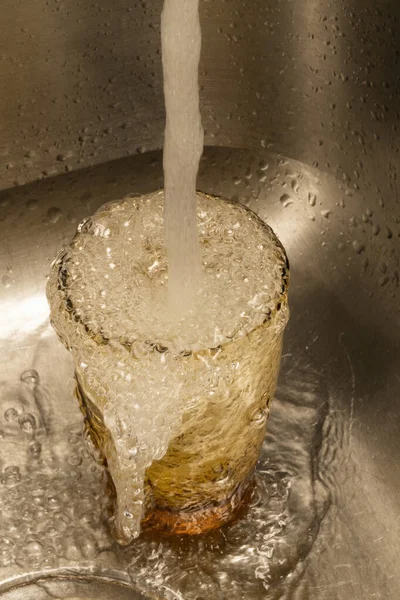 Vertical format image of water overflowing a drinking glass sitting in a stainless steel sink.