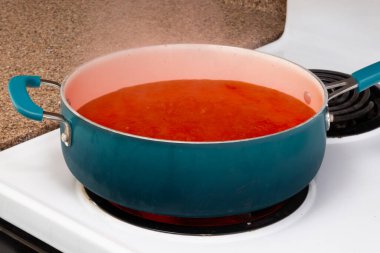 Horizontal close-up shot of a blue pot on a stove top holding steaming tomato soup.  