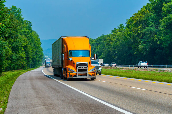 Horizontal shot of an orange eighteen wheeler in traffic on an interstate highway.