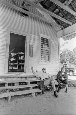 Makedonya, Arkansas, ABD 23 Haziran 2024: 1970 'lerin eski bir taşra mağazasının önünde oturup konuşan iki adamın yatay çekimi.
