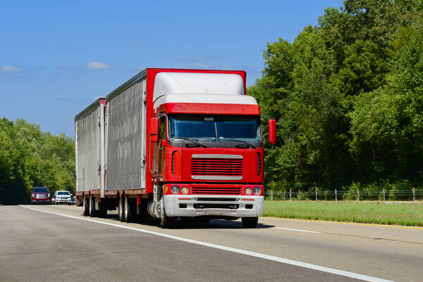 Horizontal shot of a red cab-over semi eighteen-wheeler with dual trailers.  Heat waves rising from the hot asphalt creates a blurring effect on background trucks and foreground pavement.