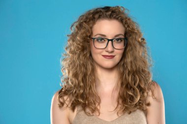 Kıvırcık saçlı, gözlüklü, mavi stüdyo arka planında gülümseyen çekici sarışın kadın..