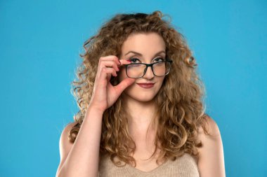 Kıvırcık saçlı, çekici sarışın kadın mavi arka planda gözlüklü poz veriyor..