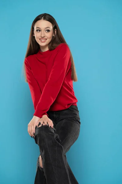 Cute smiling girl in red blouse and black jeans isolated on blue studio background