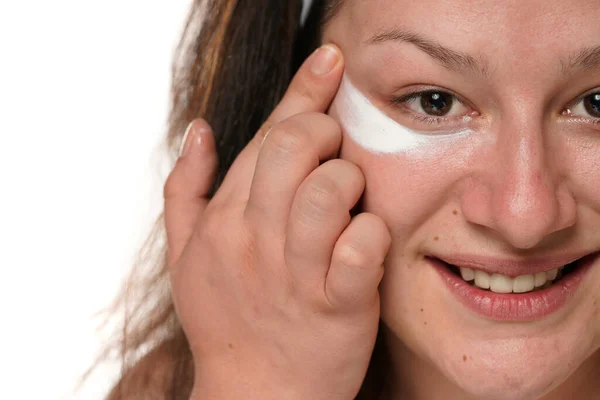 portrait of a young smiling woman chubby applying cosmetic product under her eyes on a white studio background.