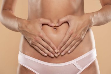 İç çamaşırlı, güzel, zayıf bir kadının göbeğine yakın ellerinde kalp şeklindeki ellerle bej renkli stüdyo arka planında poz verirken kısmen görüntüsü.