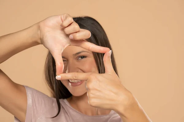 Gesturing finger frame. Beautiful young woman looking at camera and gesturing finger frame on a beige studio background.