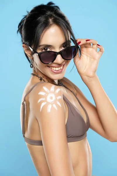 Happy Woman In Bikini With Suntan Lotion Shaped Sun On Her Shoulder, On A Blue Studio Background.