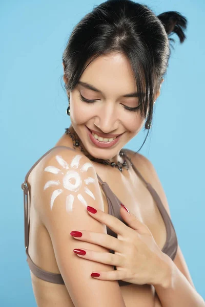 Happy Woman In Bikini With Suntan Lotion Shaped Sun On Her Shoulder, On A Blue Studio Background.
