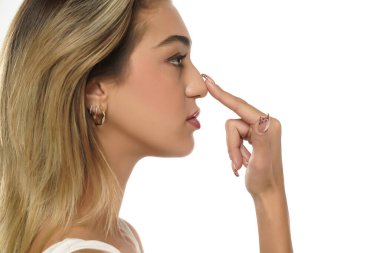 Genç bir kadın, beyaz stüdyo arka planında parmağıyla burnuna dokunuyor.