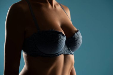 Close up on woman torso in the shadow. Blue bra and natural breasts cleavage on a blue studio background clipart