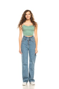 Yeşil gömlek ve mavi jeansi giyen gülümseyen genç bir kadının izole edilmiş beyaz stüdyo arka planında dikilirken tam boy fotoğrafı..