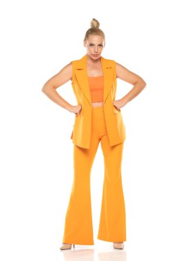 Beyaz stüdyo arka planında poz veren sarı kolsuz ceketli ve pantolonlu, orta yaşlı, sinirli bir kadın.