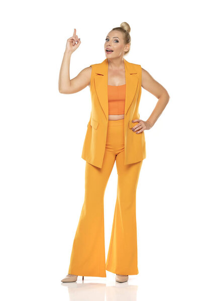 Middle aged smiling senior woman in yellow sleevless jacket and trousers pointing up on a white studio background