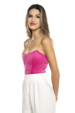 Beyaz pantolonlu, pembe korseli, beyaz stüdyo arka planında gülümseyen genç bir kadın.