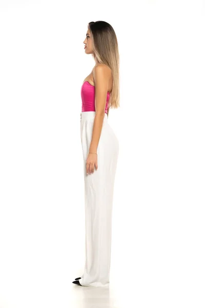 一个身穿白色长裤和粉色紧身衣的年轻现代女性在白色摄影棚背景下的侧面照片 — 图库照片