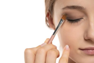 Beyaz stüdyo arka planında fırçayla göz farı süren genç bir kadının yarım portresi.