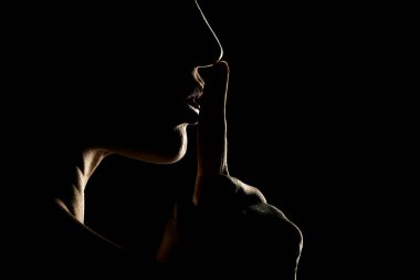 Silhouette of unknown woman with the face in the shadow holding finger on her lips on a black studio background clipart