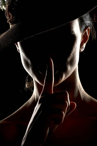 Silhouette of unknown woman with a hat, and the face in the shadow holding finger on her lips on a black studio background