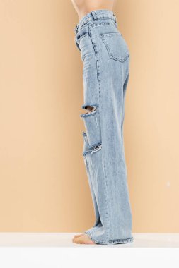 Bej renkli stüdyo arka planında yırtık kot giyen bir kadının bacakları. Yan, profil görünümü.