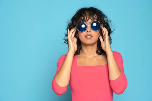 Młoda Egzotyczna Brunetka Różowej Bluzce Pozująca Okularami Przeciwsłonecznymi Niebieskim Tle — Zdjęcie stockowe