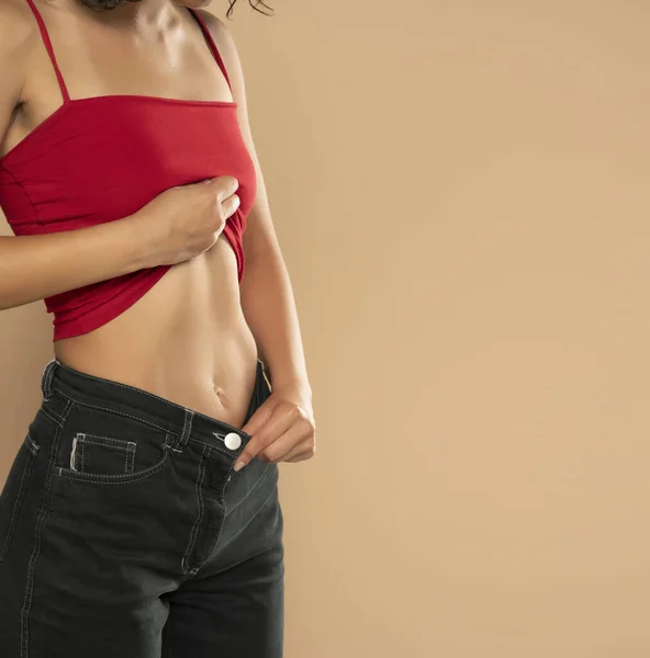穿着红色无袖衬衫和牛仔裤的年轻女子在米色的工作室背景上露出扁平的腹部 — 图库照片