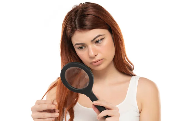 忧心忡忡的红头发女人拿着放大镜在白色的工作室背景下检查她的头发 — 图库照片