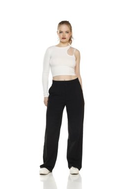 Beyaz stüdyo arka planında beyaz bir bluz ve pantolon giyen sarışın genç bir kadının ön görüntüsü.