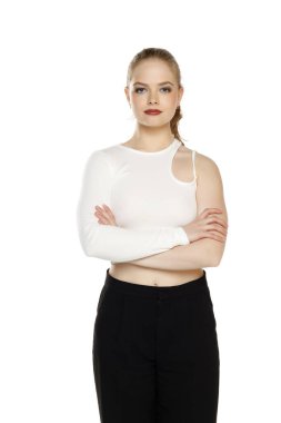 Beyaz stüdyo arka planında kolları katlanmış beyaz bluz ve pantolonlu genç sarışın kadının ön görüntüsü.