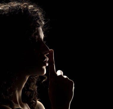 Bilinmeyen bir kadının silueti. Yüzü gölgede, siyah bir stüdyo arka planında parmaklarını dudaklarında tutuyor.