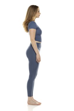 Tüm vücut, yan görüş spor giyimli genç kadın beyaz stüdyo arka planında poz veriyor.