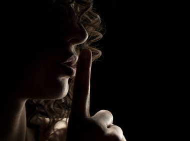 Bilinmeyen bir kadının silueti. Yüzü gölgede, siyah bir stüdyo arka planında parmaklarını dudaklarında tutuyor.