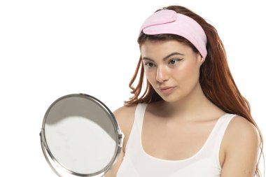 Saç bandı takmış genç kızıl saçlı güzel bir kadın beyaz bir stüdyoda aynada kendine bakıyor.